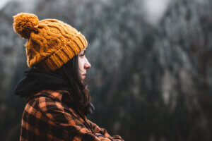 Side view of thoughtful young woman wearing plaid shirt and yellow winter hat looking at view in cold weather conditions. Travel, outdoor and cold weather concept. - mental preparation for the holidays