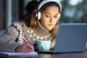 Young woman wearing headphones with laptop taking notes on notebook in winter