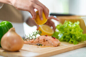 Close up of woman's hand squeezing lemon juice on a piece of salmon steak in the kitchen for dinner at home. Happy female enjoy cooking and eating healthy food on holiday vacation
