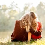 two young women sitting on grass hugging rear view